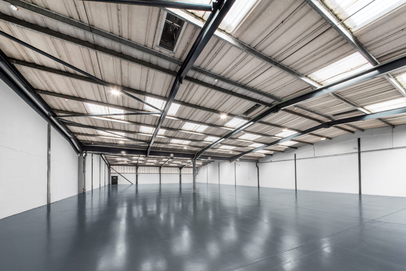Large empty warehouse interior with high ceilings, exposed beams, white walls, and a polished concrete floor. Bright overhead lighting illuminates the spacious, open area.