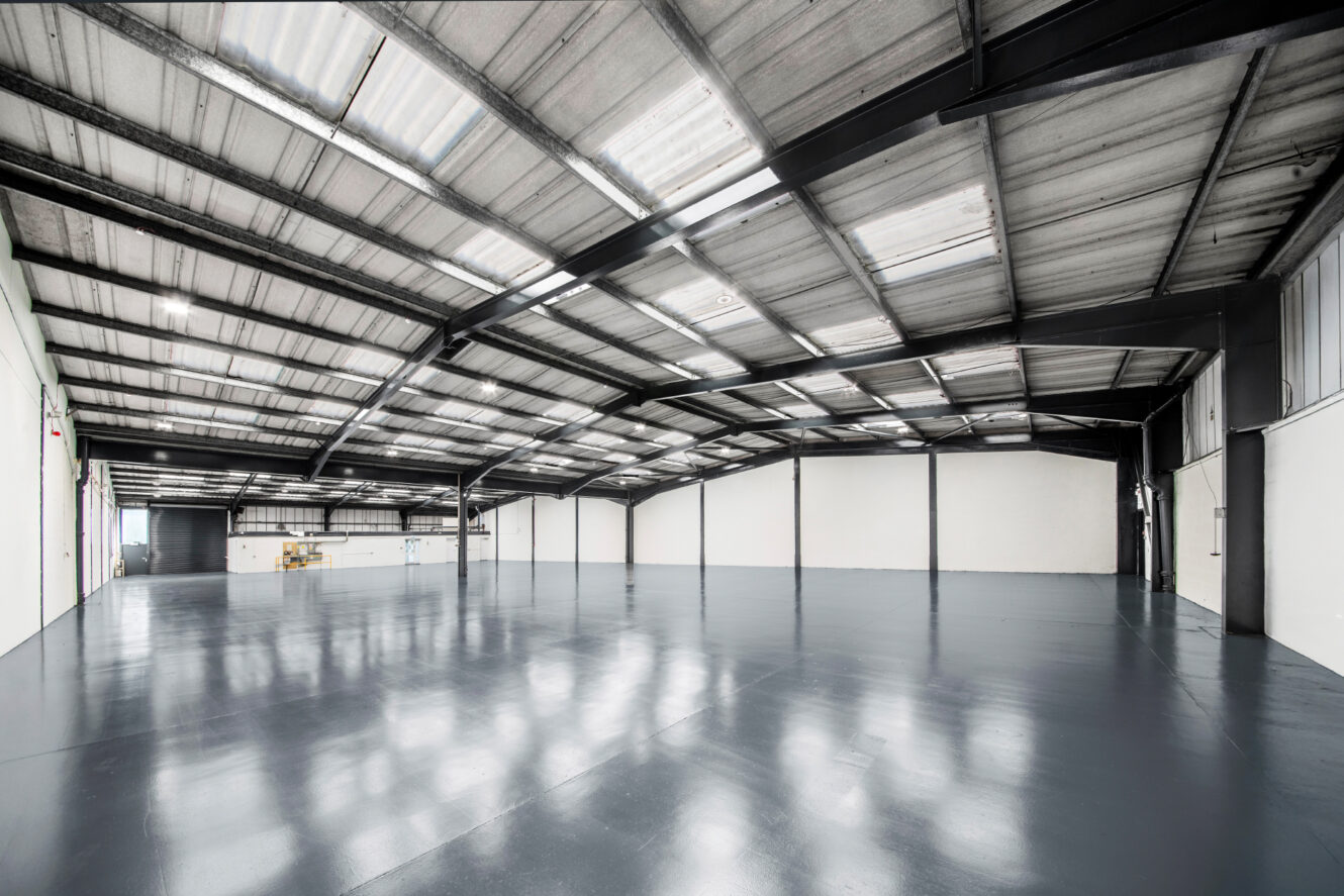 Large, empty industrial warehouse with a polished concrete floor, white walls, and a high metal ceiling with skylights.