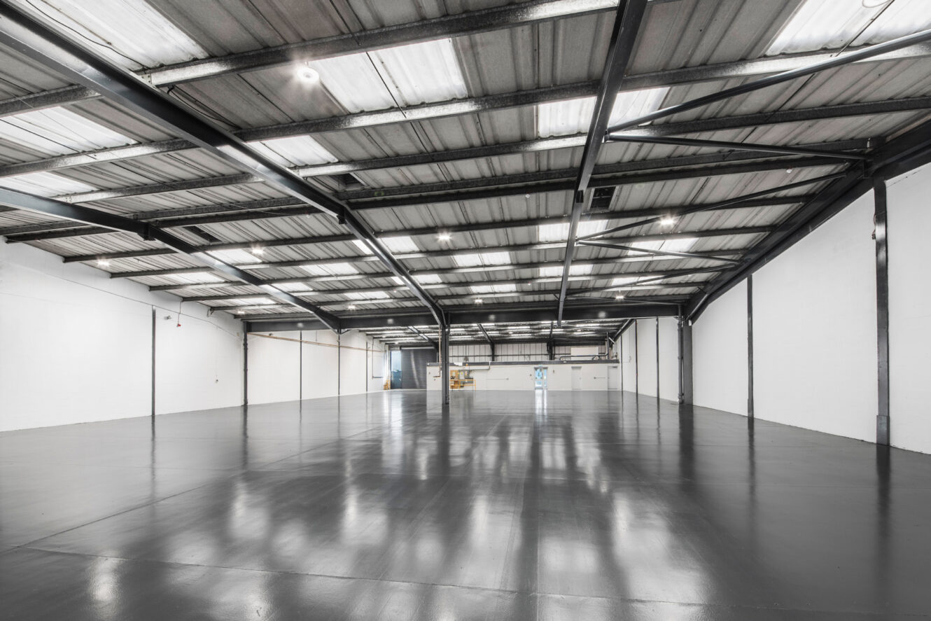 Spacious, empty warehouse with polished concrete floors, high ceilings, exposed beams, and fluorescent lighting. Boxes are stacked near the far wall.