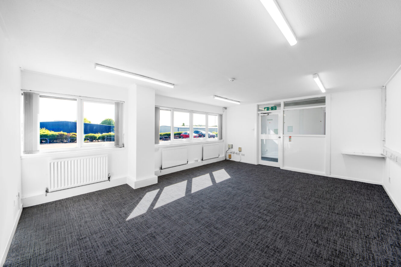 A bright, empty office room with white walls, large windows, grey carpet, ceiling lights, and visible radiators, with sunlight streaming in through the windows.