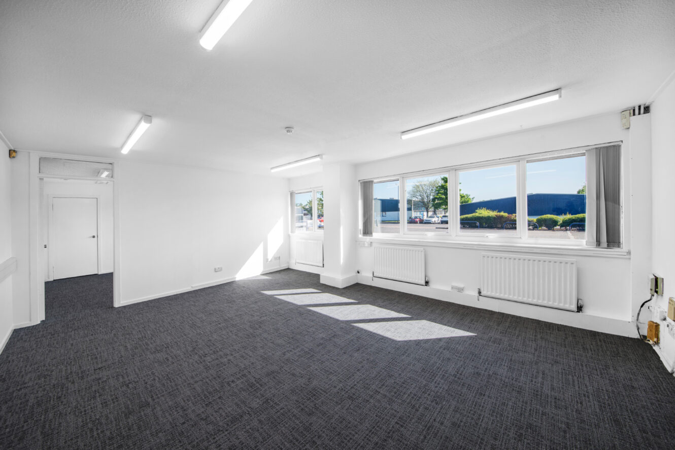 Empty office room with white walls, grey carpet, fluorescent lighting, large windows, and a door at the back.