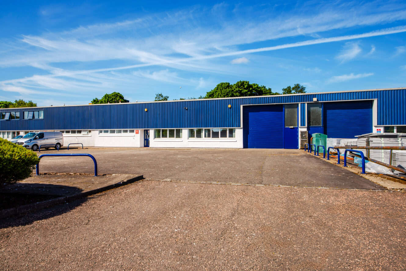 Single-story industrial warehouse with blue doors and a blue facade, surrounded by an asphalt lot and a few parked cars under a clear sky.