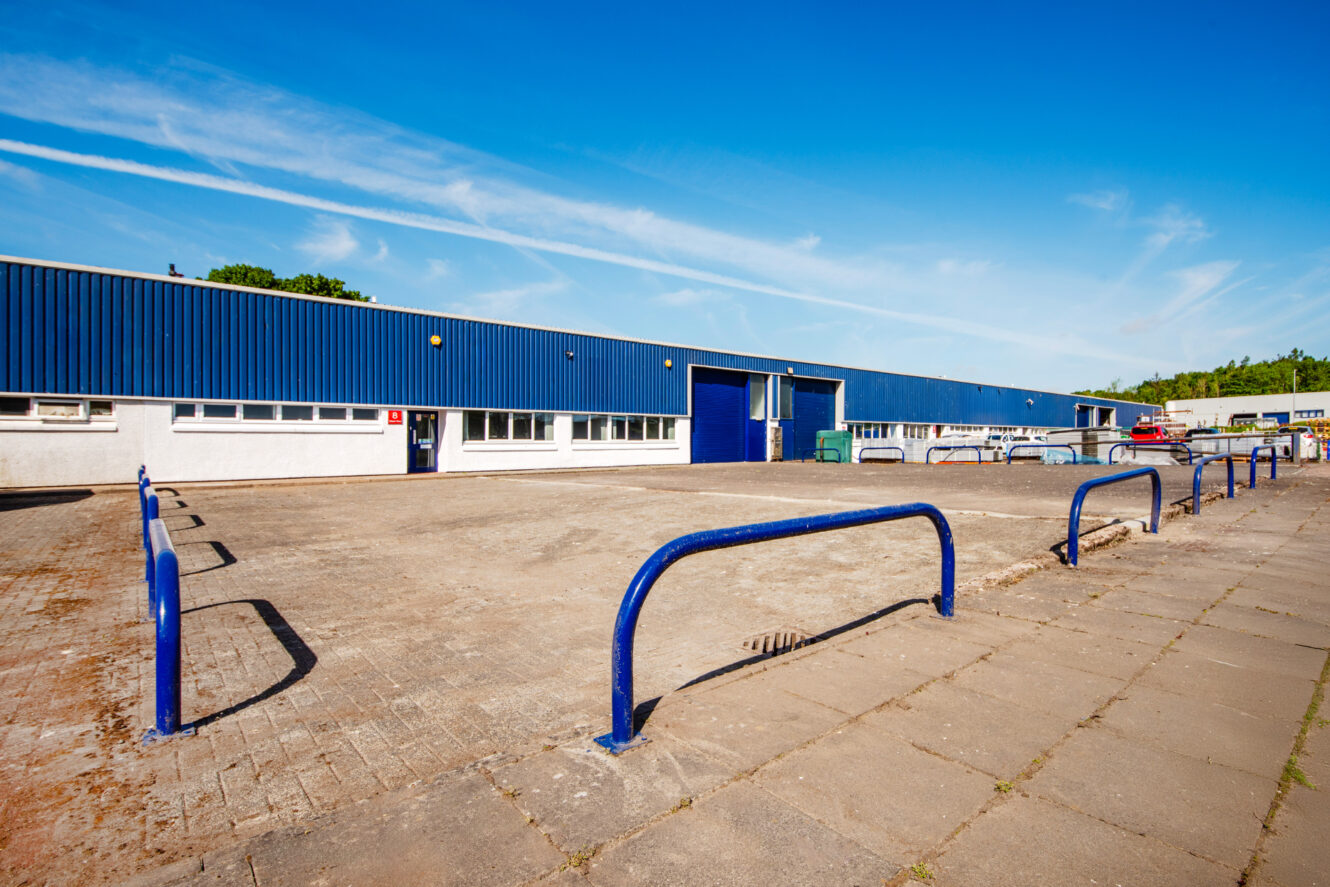 Industrial warehouse with blue siding and roller door, surrounded by blue safety barriers and a paved area, under a clear blue sky with contrails.