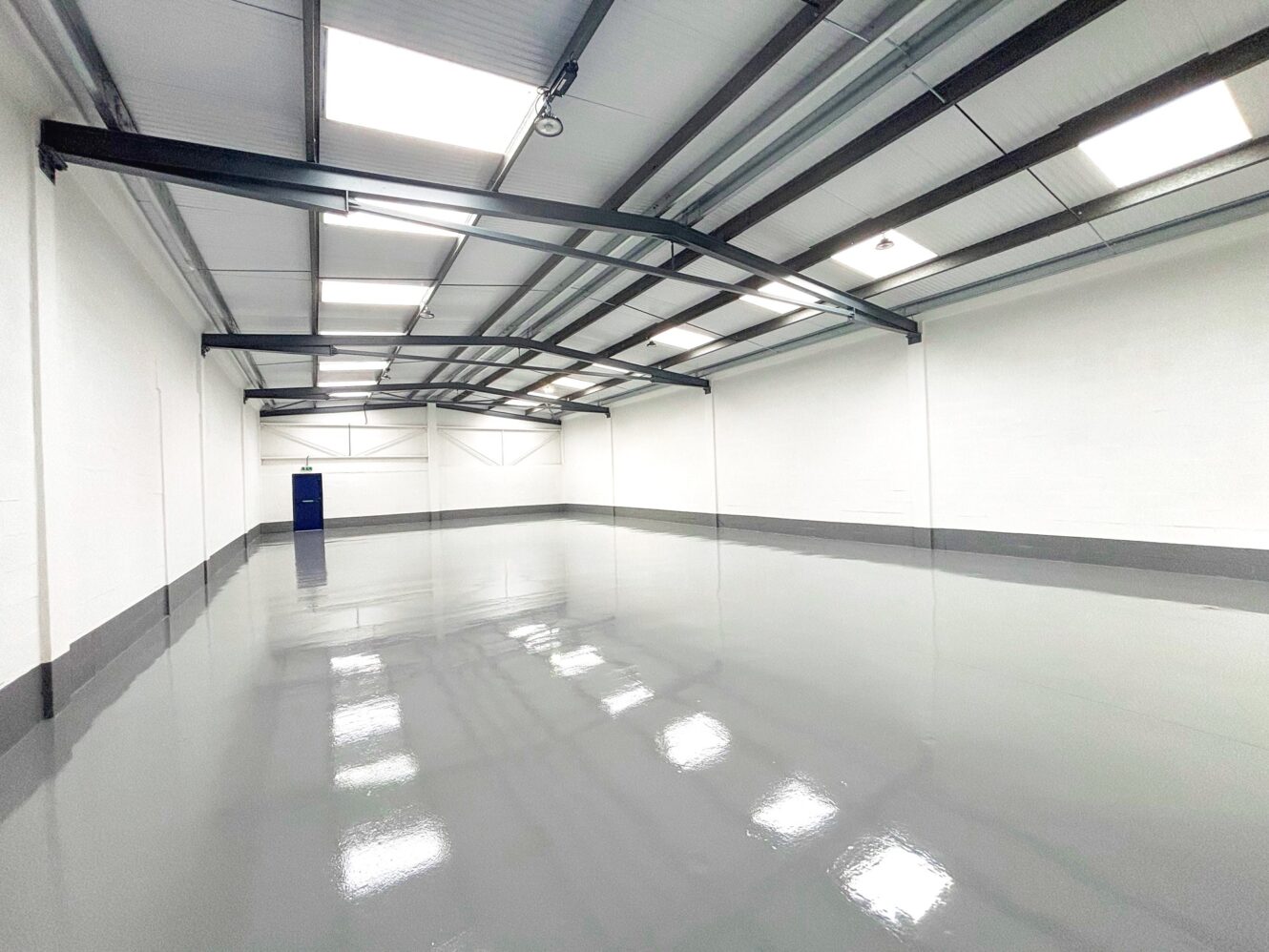 Empty industrial warehouse with white walls, a polished gray floor, exposed ceiling beams, and fluorescent lights. A blue door is visible at the far end.