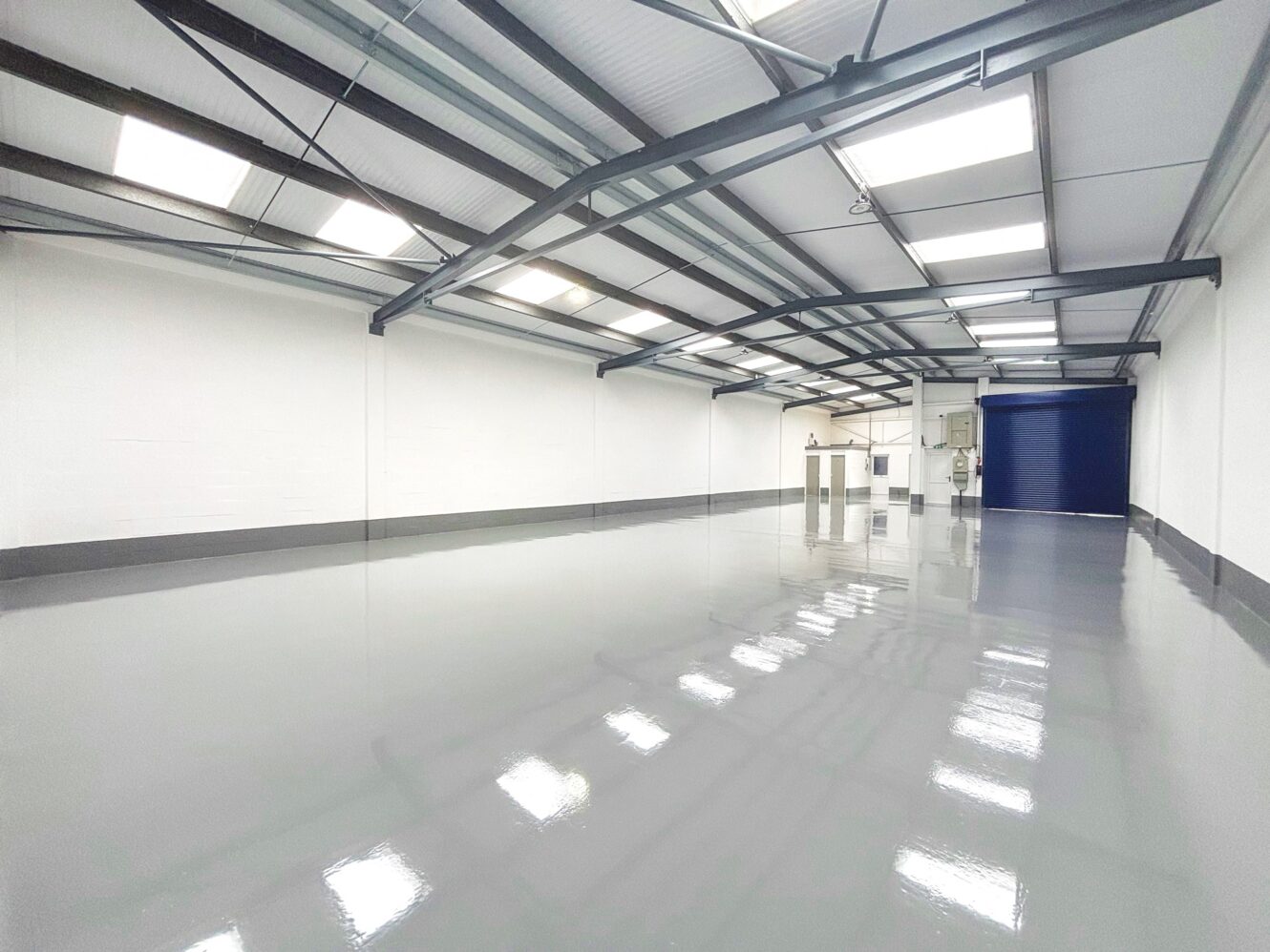 An empty industrial warehouse with white walls, high ceilings, metal beams, and a polished gray floor under fluorescent lighting.