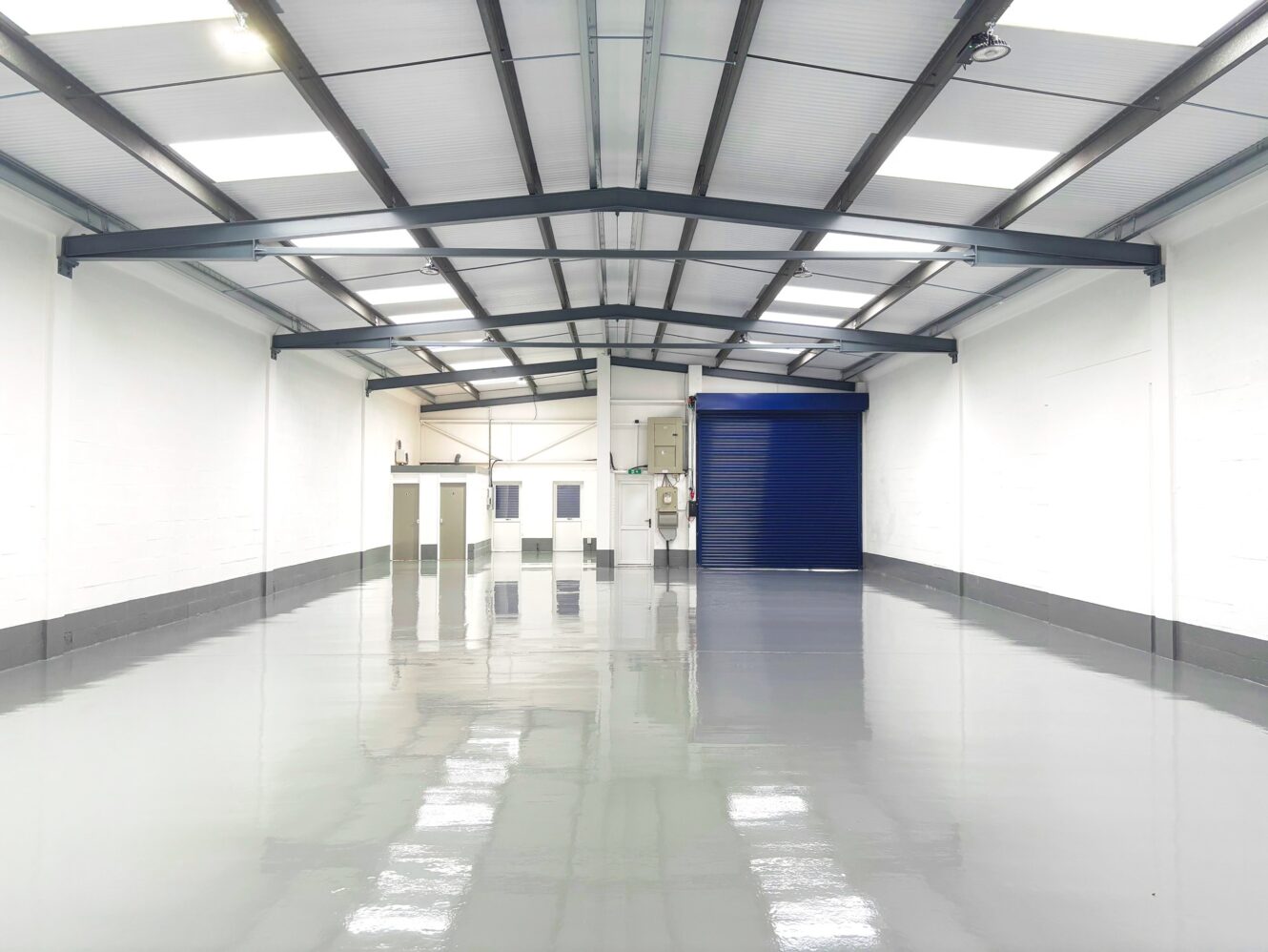 A clean, empty warehouse with white walls, a polished gray floor, and a blue roller shutter door at the far end. Ceiling lights and steel beams are visible overhead.