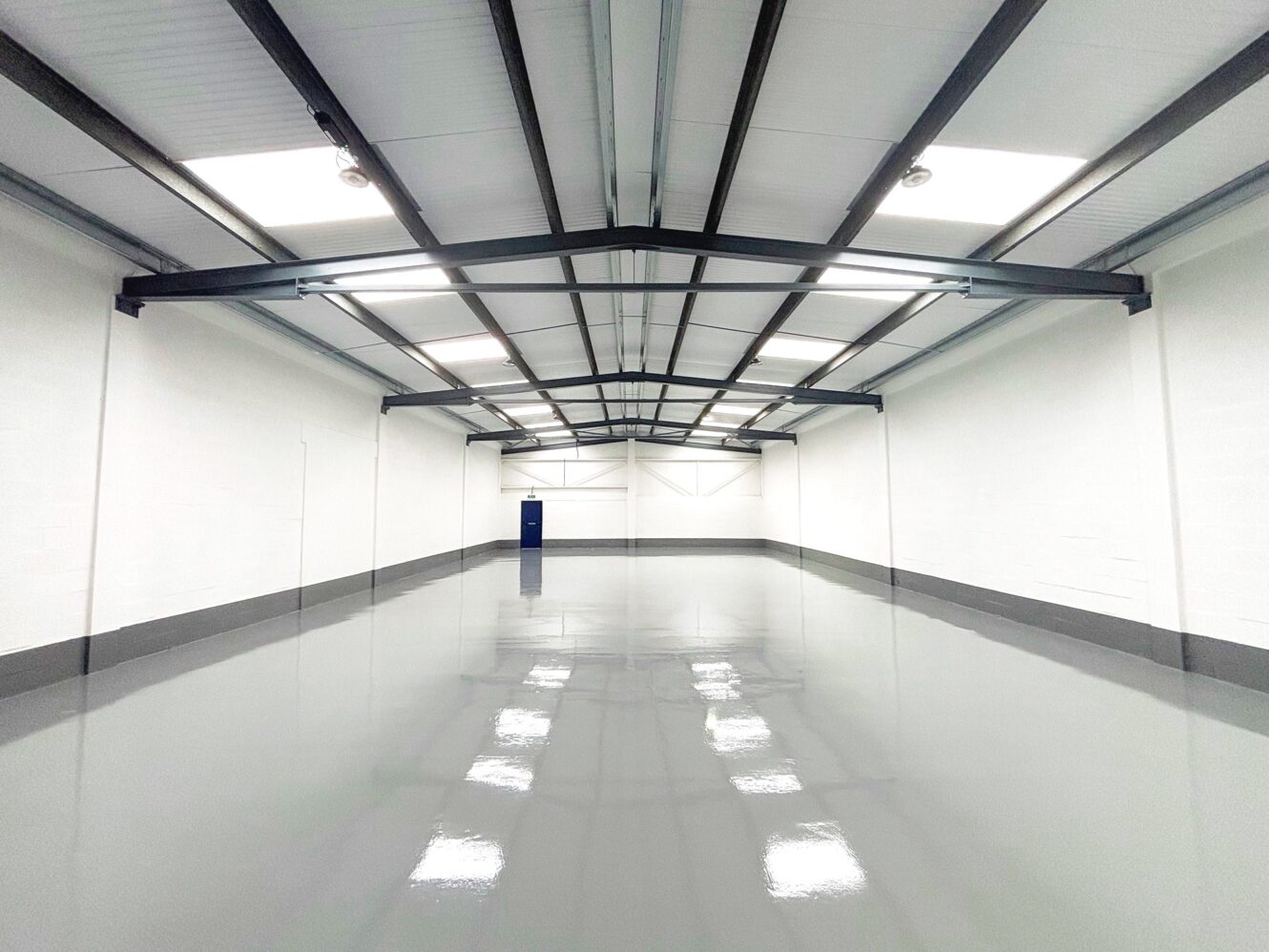 Empty industrial warehouse with white walls, a grey polished floor, exposed steel beams, and a single blue door at the far end.