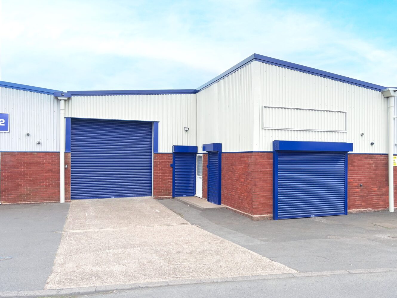 A small industrial warehouse with metal siding, red brick lower walls, and blue roller shutter doors, situated on an empty lot under a clear sky.