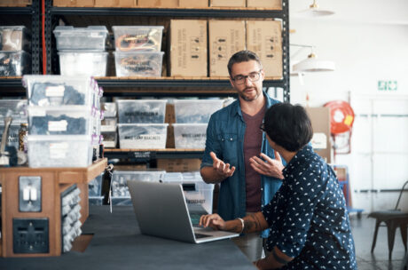 Two people discuss work at a counter in a warehouse, with a laptop open in front of them and shelves of organized boxes and bins in the background.
