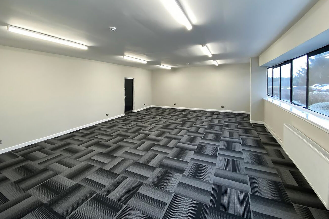 Empty office room with patterned carpet tiles, white walls, fluorescent ceiling lights, a black door, and large windows along one side letting in daylight.