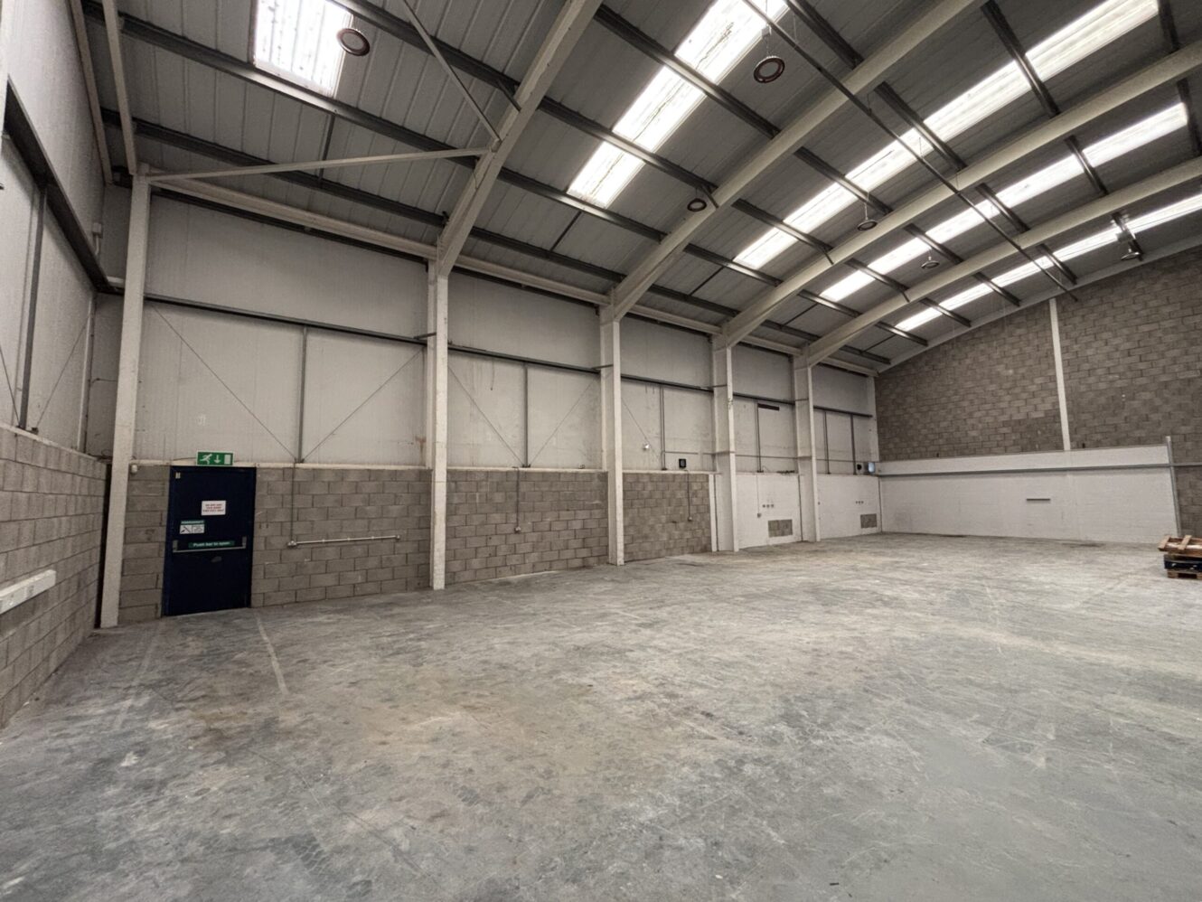 Empty industrial warehouse with high ceilings, grey concrete floors, brick walls, fluorescent lighting, and a single dark door on the left.