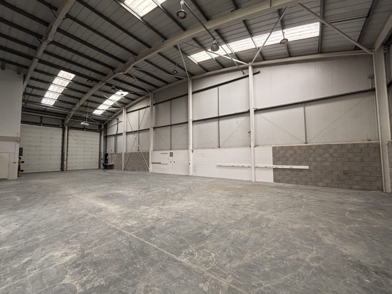 An empty industrial warehouse interior with a high ceiling, large windows, concrete floor, and a closed roller shutter door.