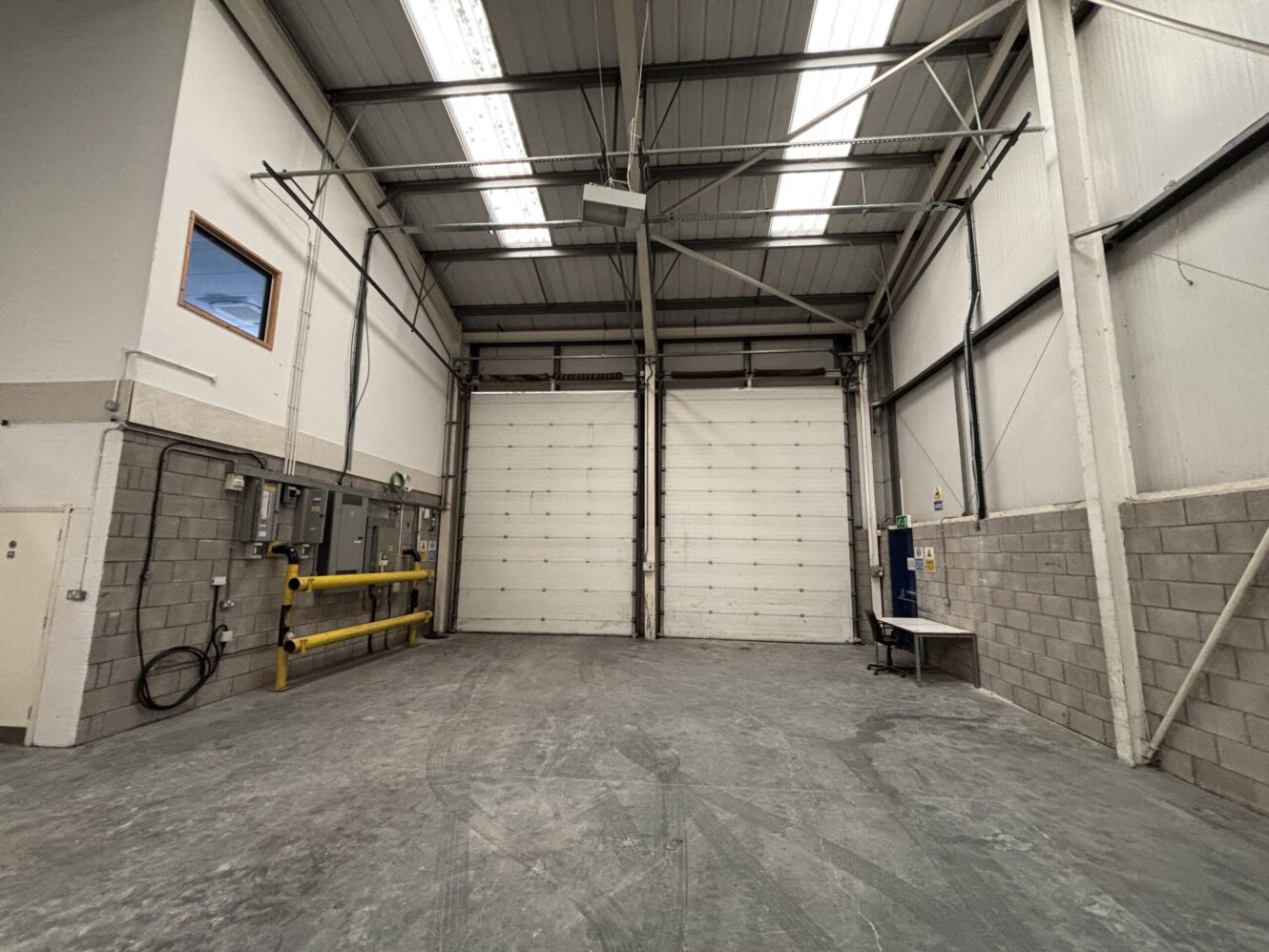 An empty industrial warehouse with a large closed roller door, concrete floor, gray block walls, and exposed ceiling beams.
