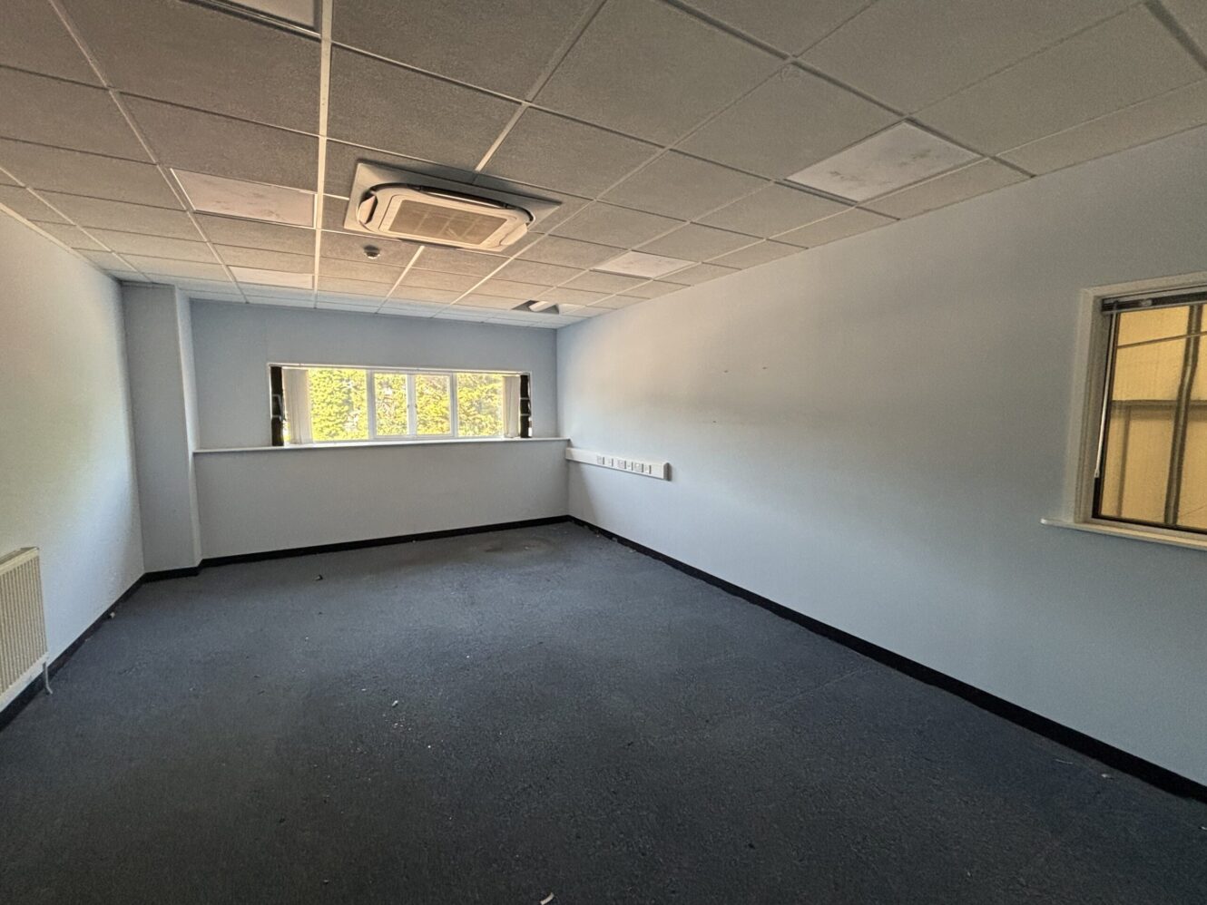 Empty office room with gray carpet, white walls, three windows, ceiling tiles, an air conditioning unit, and a small window into another room.