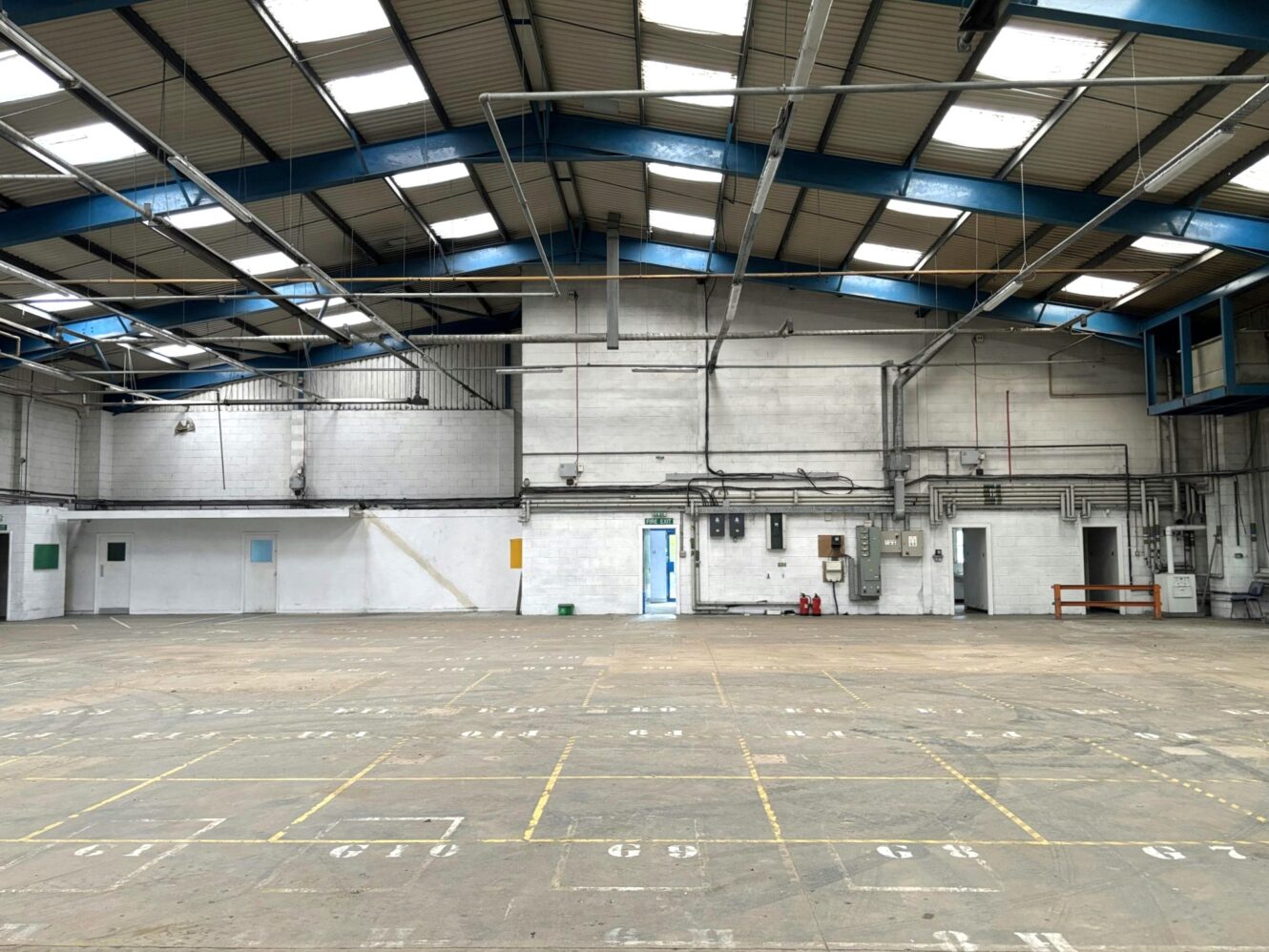An empty industrial warehouse with a high ceiling, exposed beams, marked floors, and utility panels on the far wall. Daylight enters through roof windows.