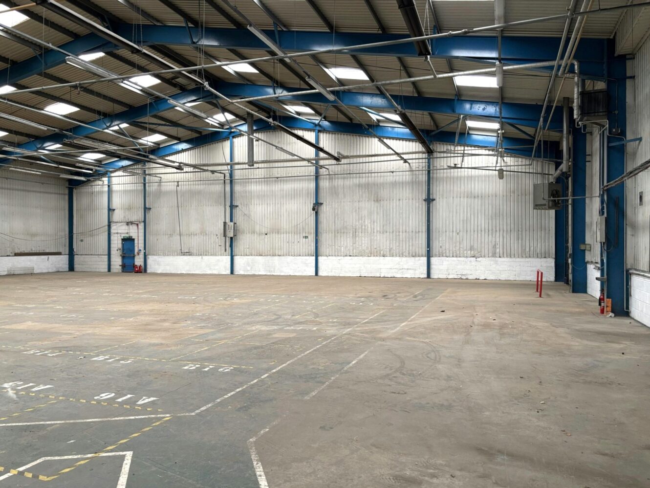 Interior of an empty industrial warehouse with concrete floor, high ceilings, blue steel beams, and white corrugated metal walls.