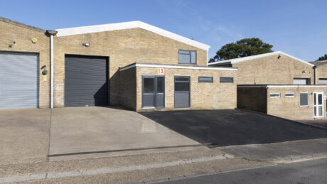 A light brown brick industrial warehouse building with a black roller shutter door, glass double doors, and a sign with the number 13 above the entrance.
