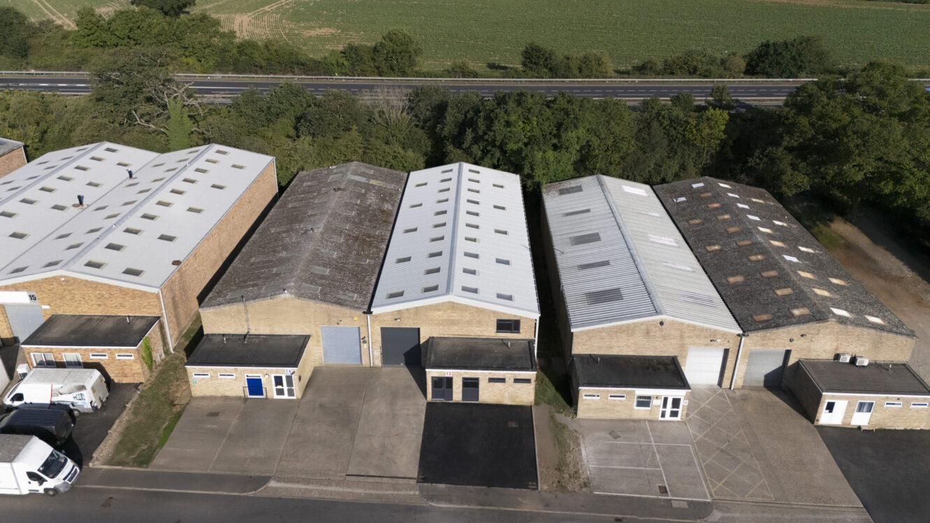 Aerial view of four industrial warehouse buildings with varying roof conditions, parking spaces, and surrounding trees near a main road and open fields.