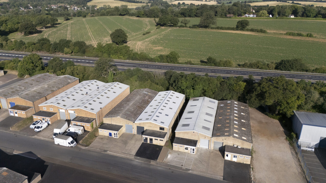 Aerial view of several industrial warehouse buildings near a road, with open fields and greenery in the background.