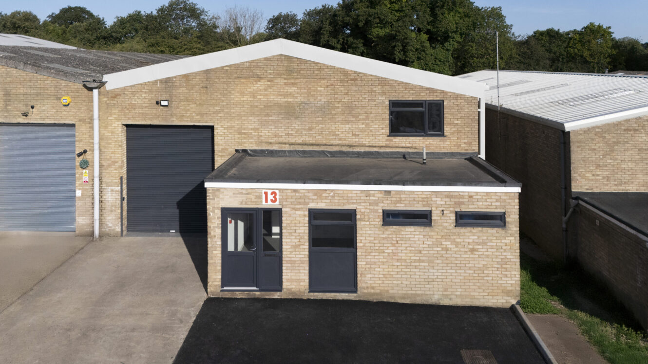 A light brown brick industrial building with a black garage door, a small office entrance, and the number 13 above the door. Trees are visible in the background.