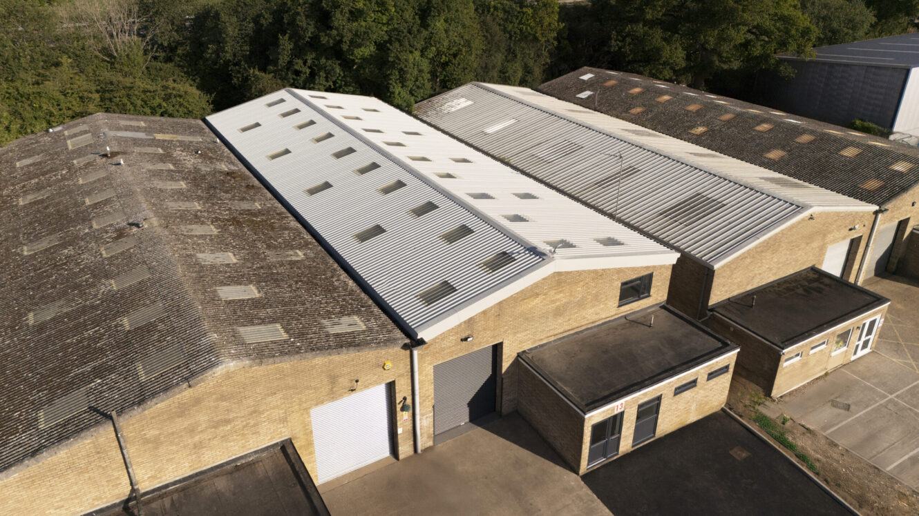 Aerial view of industrial warehouse buildings with corrugated metal roofs, skylights, and brick walls, surrounded by trees.