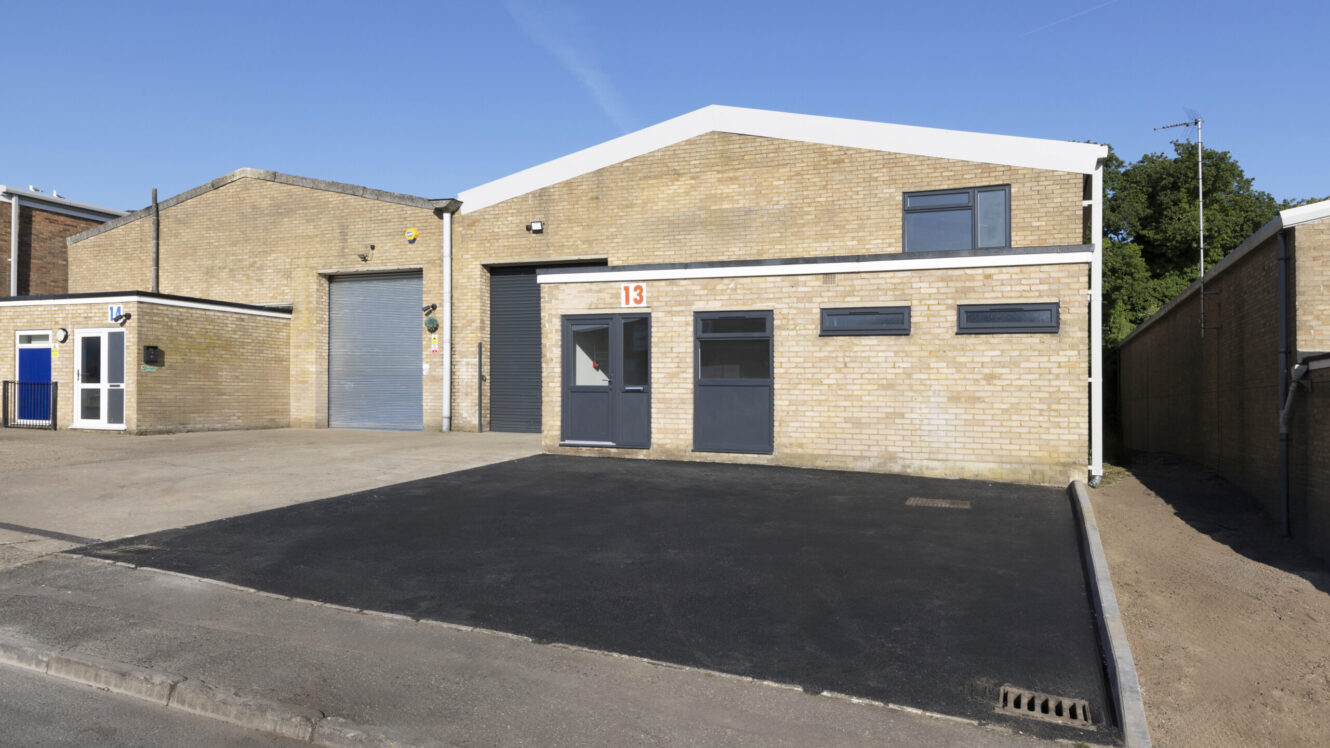 Single-story industrial warehouse with brick exterior, two dark gray doors, several windows, and a freshly paved driveway. The number 13 is displayed above the entrance.