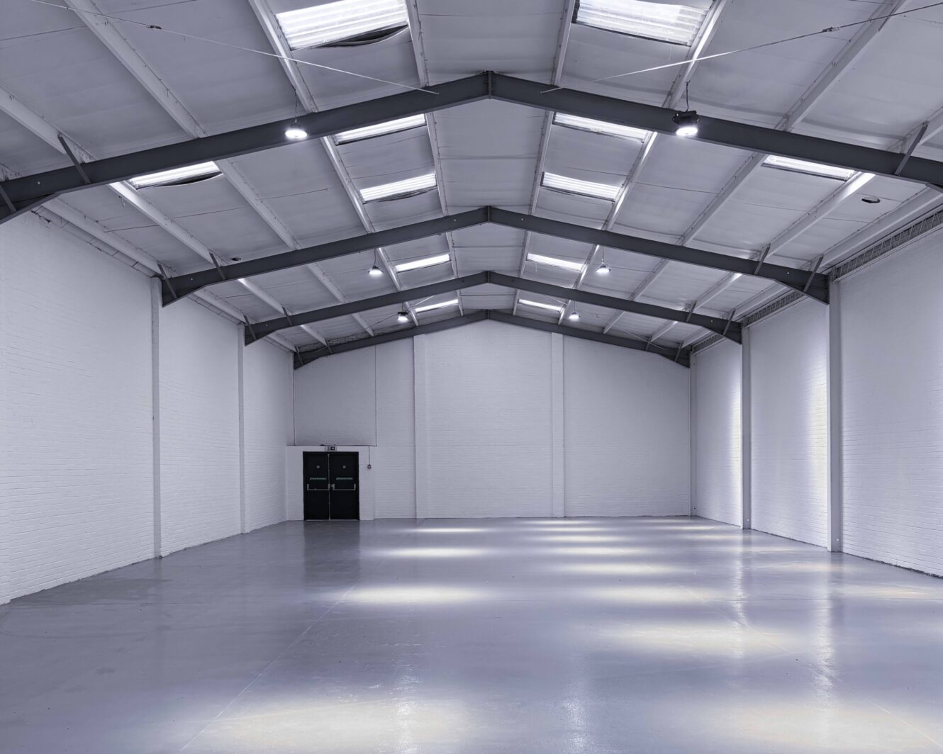 Large empty warehouse with white walls, polished floor, and high ceiling with metal beams and skylights. Double doors are visible at the far end. Bright overhead lighting.