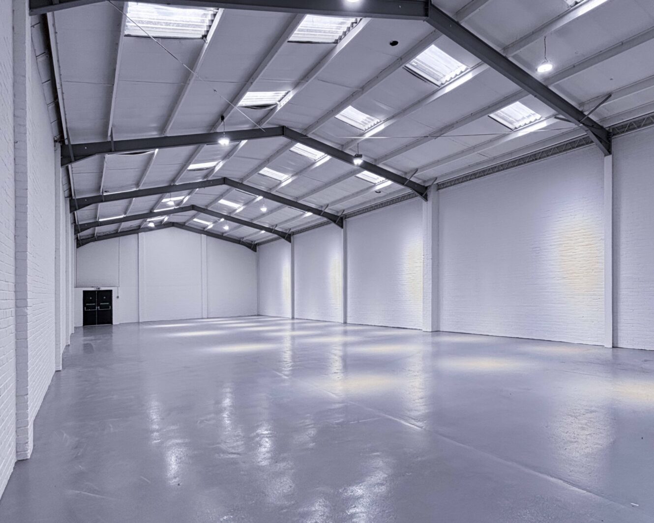 Large empty warehouse with white brick walls, gray concrete floor, and a high ceiling with skylights and exposed metal beams. Double doors are visible at the far end.