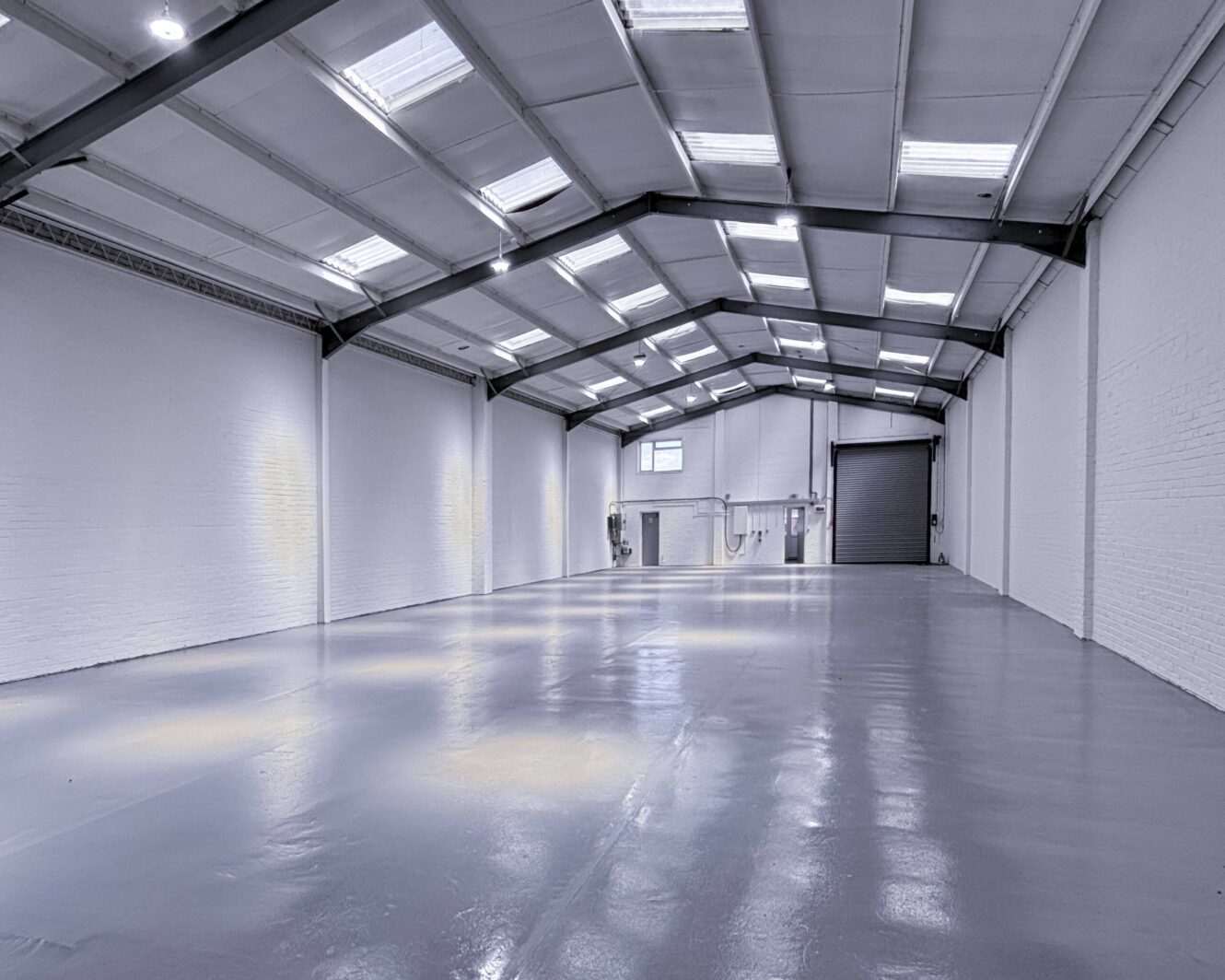 Spacious, empty warehouse with a polished floor, white walls, high ceiling, skylights, and a large roller shutter door at the far end.