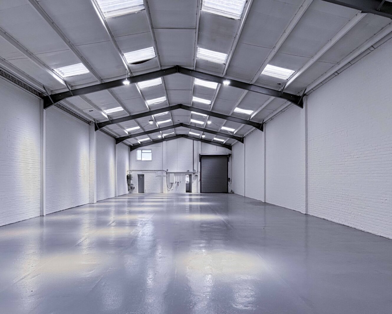 Interior of an empty, modern warehouse with white walls, a polished concrete floor, skylights, and a large roller door at the far end.