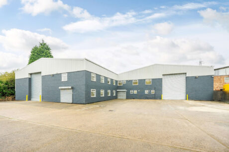 Industrial warehouse building with gray exterior, large roller doors, small windows, and a spacious concrete yard under a partly cloudy sky.