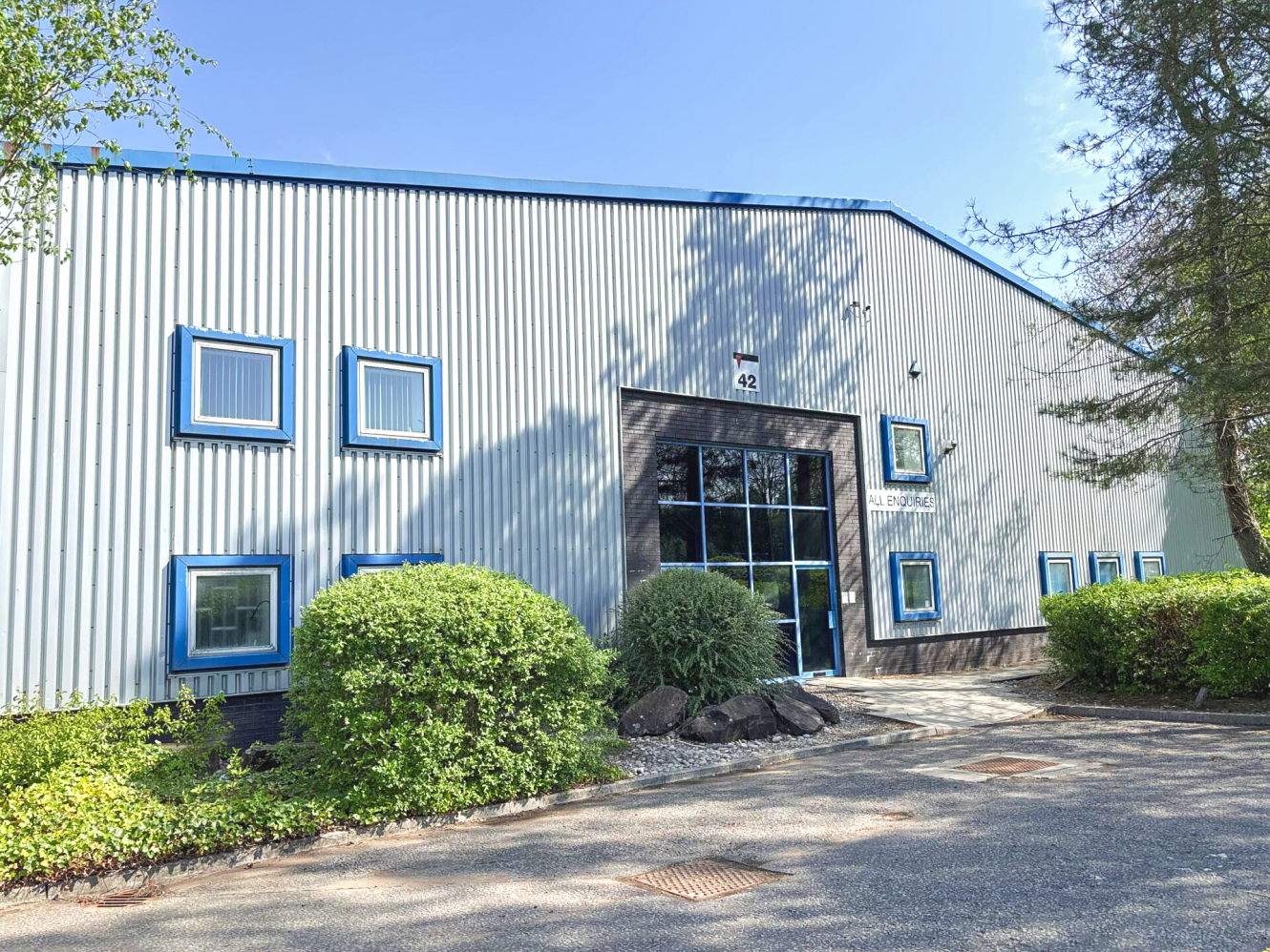 A large industrial building with a metal exterior, blue trim, multiple square windows, and glass double doors, surrounded by bushes and trees.
