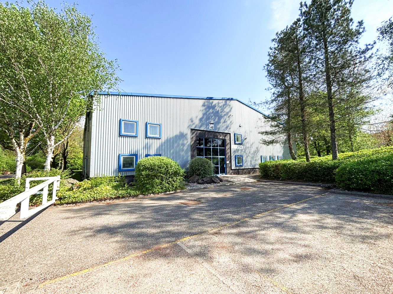 A rectangular industrial building with a metal exterior, several small square windows, and surrounding trees and bushes, viewed from an empty parking lot.