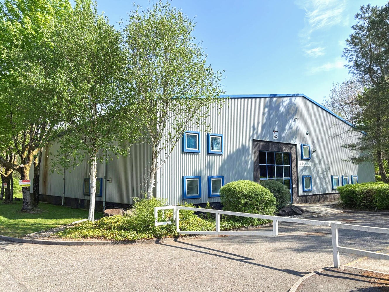 A light gray industrial building with blue-trimmed windows and a large entrance, surrounded by trees and greenery under a partly cloudy sky.