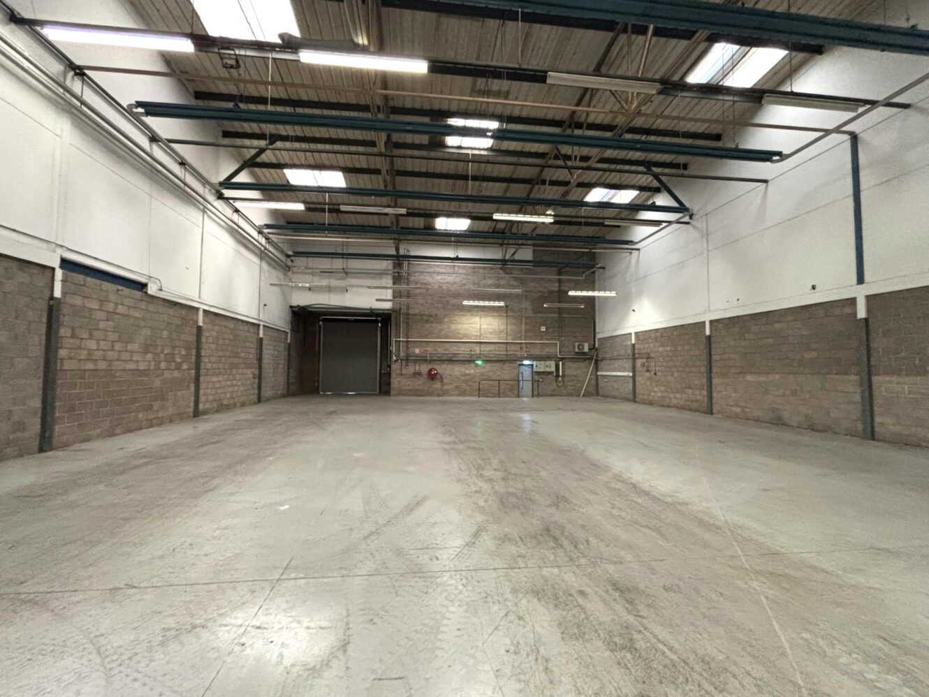 Large, empty warehouse space with concrete floors, high ceilings, exposed beams, brick walls, and a raised loading door at the far end.
