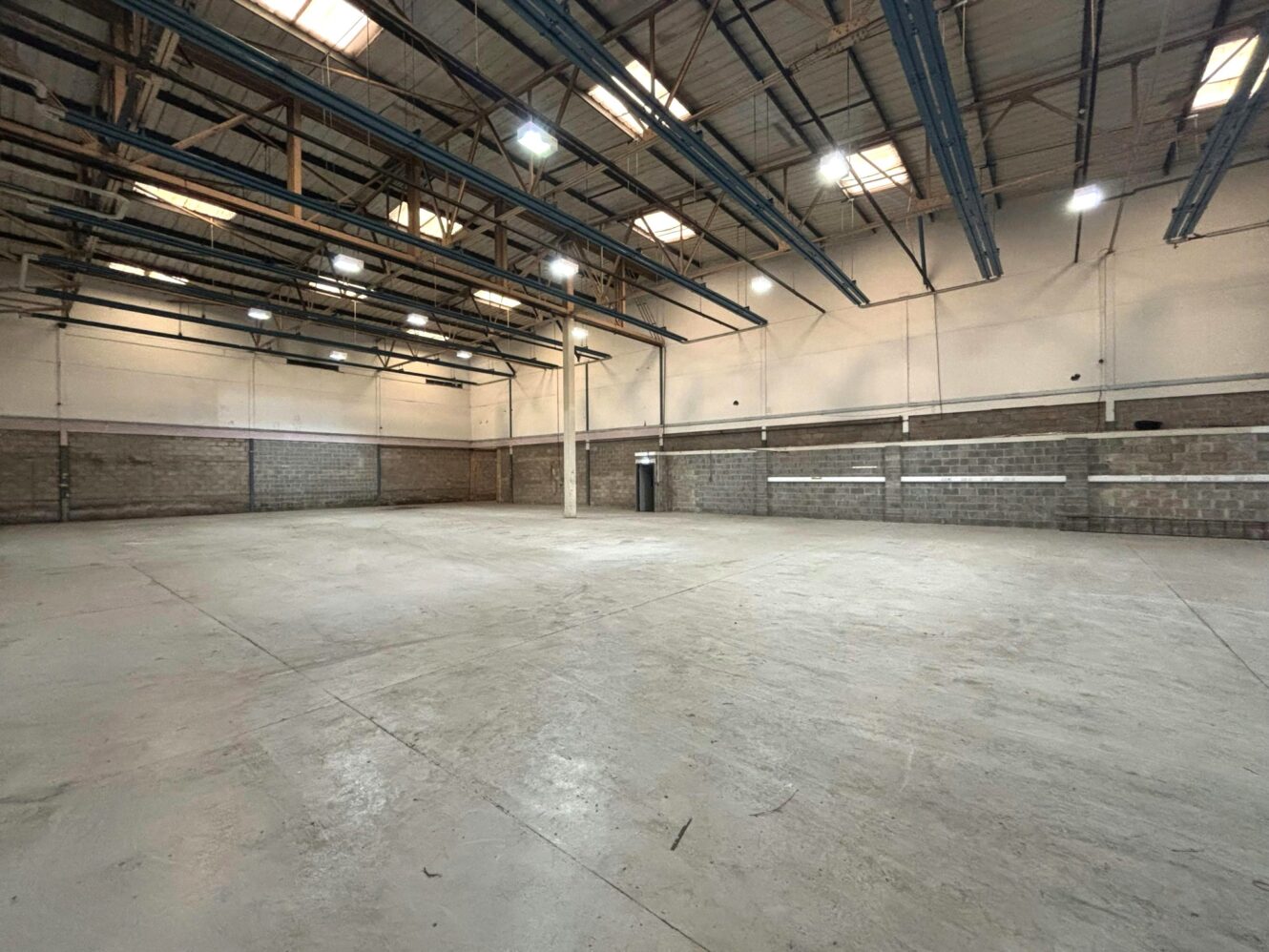 Large, empty industrial warehouse with concrete floors, high ceilings, exposed beams, skylights, and cinder block walls.