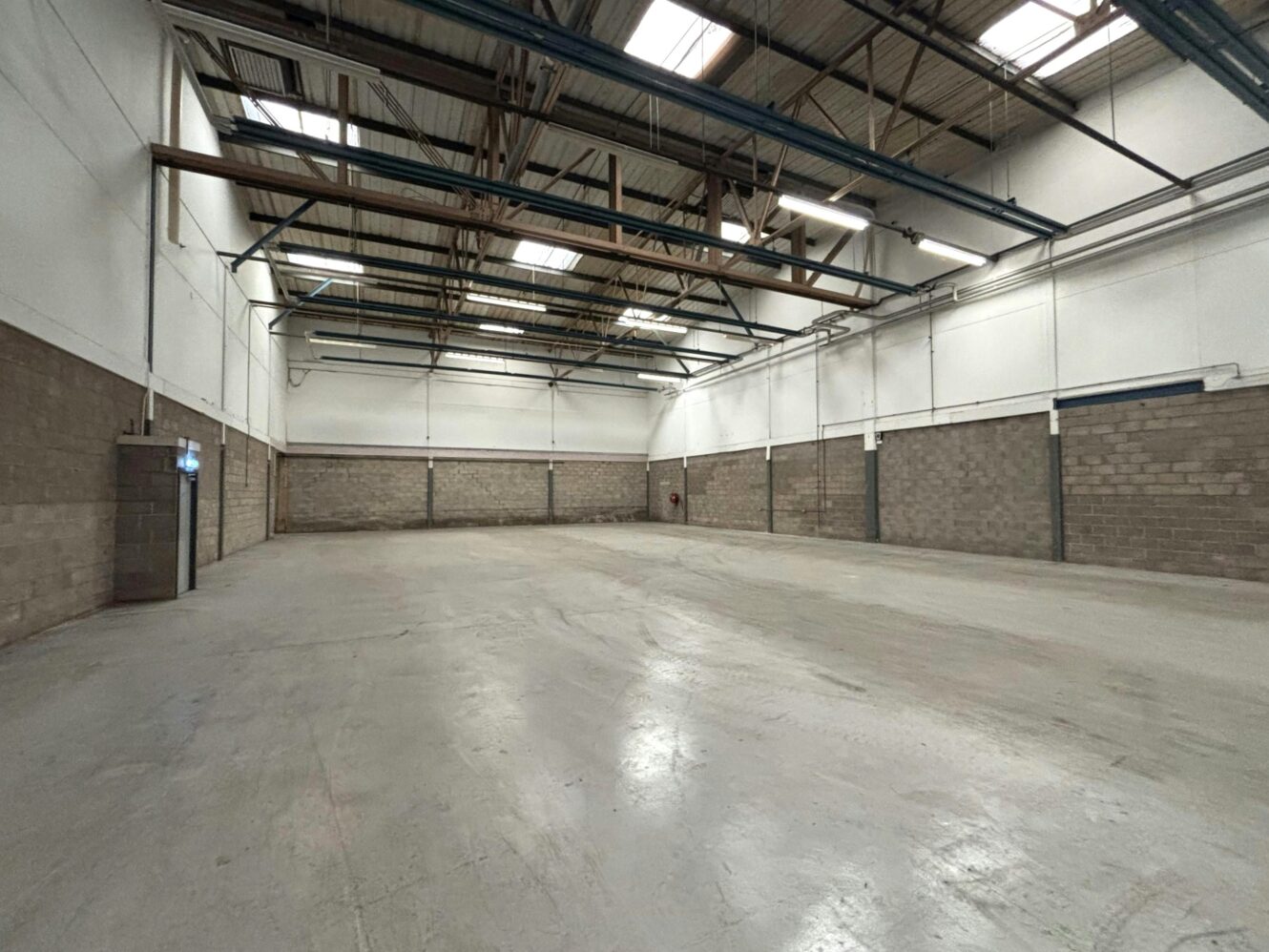 An empty industrial warehouse with a high ceiling, exposed beams, concrete floors, and brick lower walls, illuminated by overhead lights and skylights.