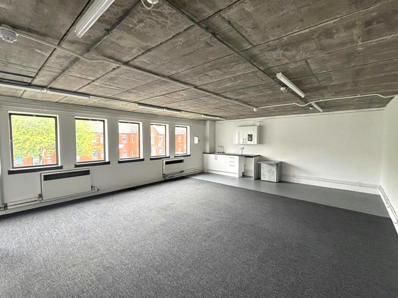 Unfurnished office space with carpeted floor, exposed concrete ceiling, large windows, and a small kitchenette in the corner.