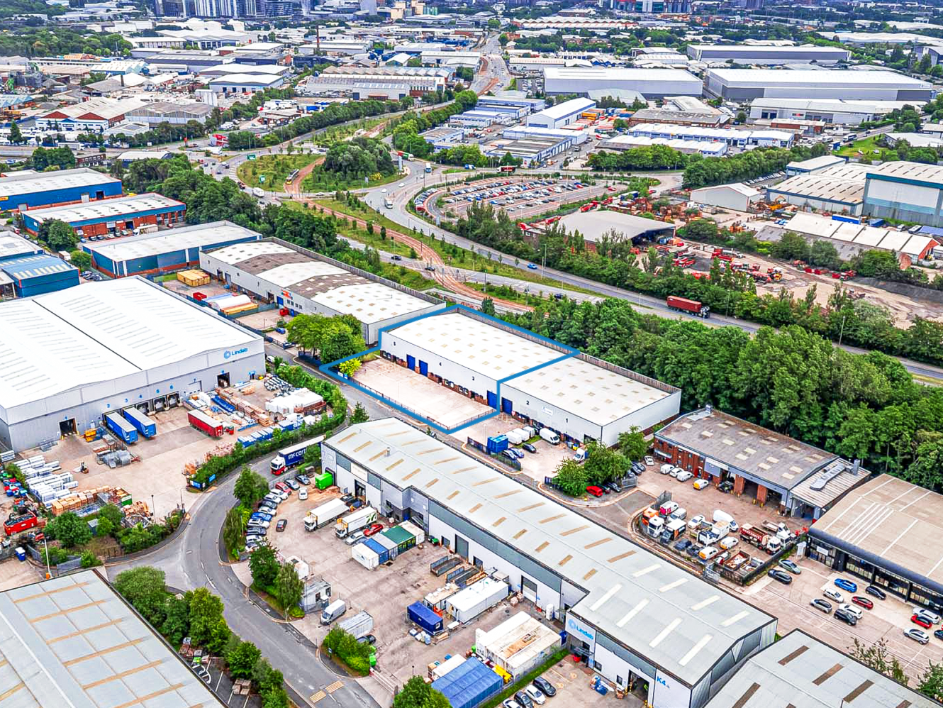 Aerial view of an industrial area with large warehouse buildings, storage yards, parked vehicles, and surrounding roads and greenery.
