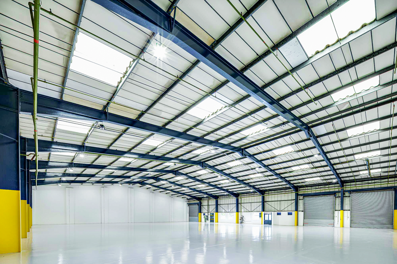 Interior of a large, empty warehouse with high ceilings, skylights, steel beams, and several large roller doors along one wall.