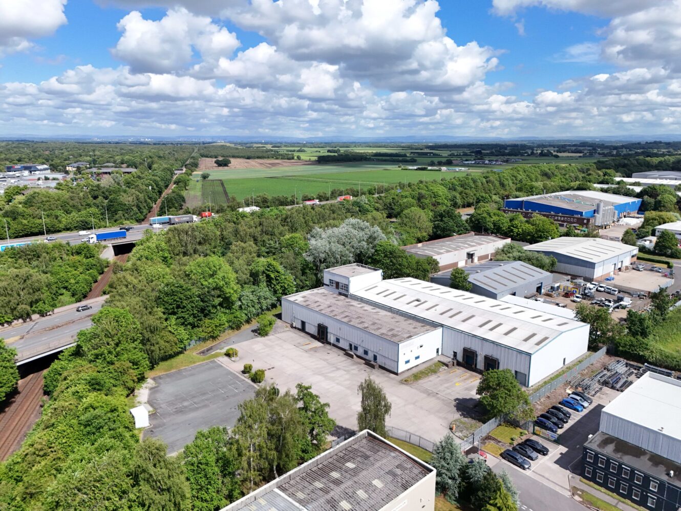 Aerial view of an industrial area with warehouses, surrounding trees, parking lots, roads, and fields in the distance under a partly cloudy sky.