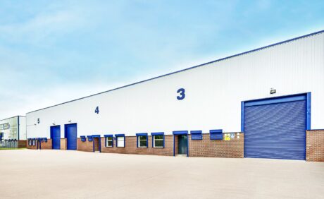 A row of blue industrial warehouse doors numbered 3, 4, and 5 on a large white and brick building with an empty concrete lot in front.