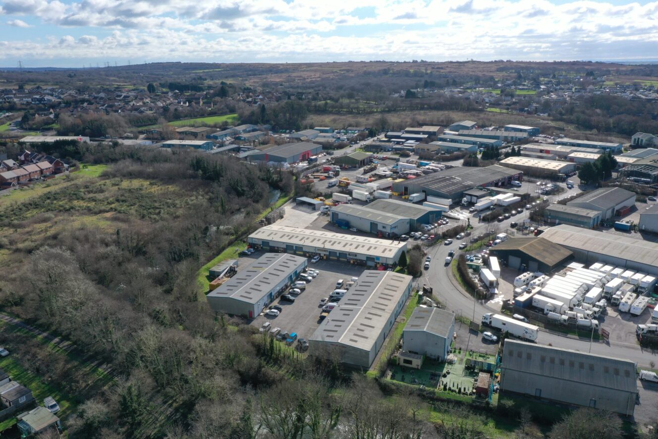 Aerial view of an industrial estate with warehouses, parked vehicles, and surrounding trees, set near a residential area and open fields under a partly cloudy sky.