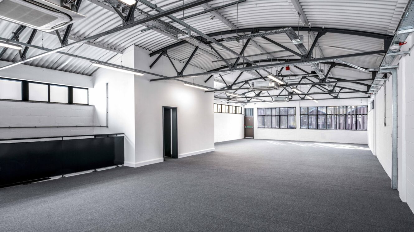 Spacious, empty industrial-style room with exposed metal ceiling beams, large windows, white walls, and gray carpeted floor.