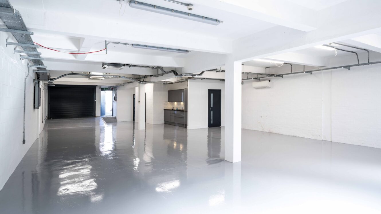 A clean, empty industrial space with white walls, exposed pipes, a polished floor, kitchen area, and a large black roller door at the back.