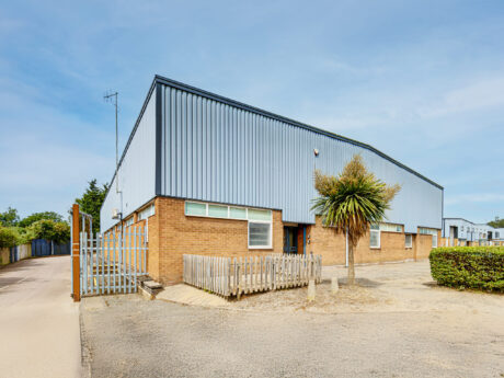 A modern industrial warehouse with brick lower walls, metal upper siding, a security gate, and a small palm tree in front.