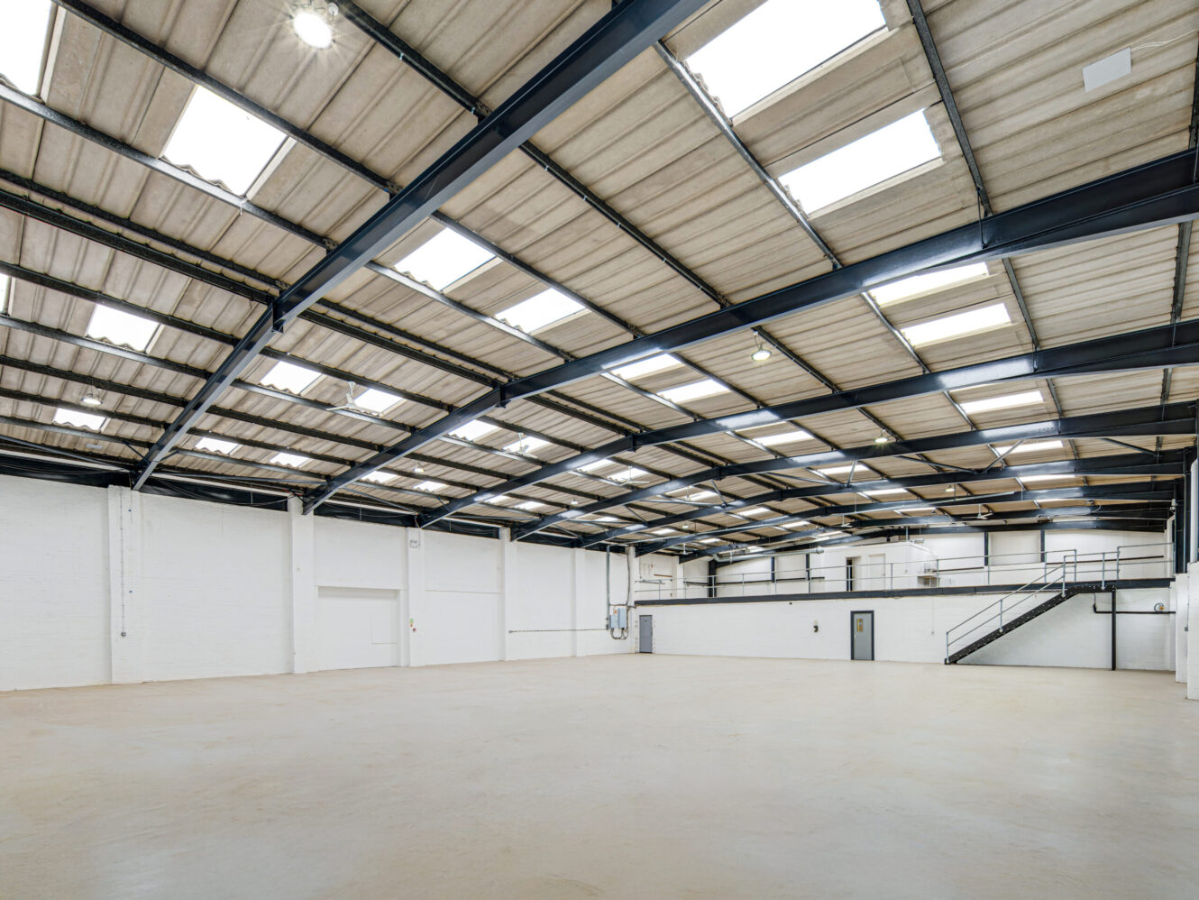 Spacious empty warehouse with high ceiling, skylights, white walls, and a mezzanine level accessed by a black staircase.