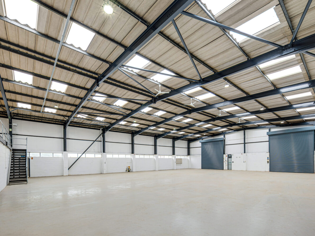 Spacious, empty warehouse with high ceilings, metal beams, skylights, white walls, concrete floor, roller shutter doors, and a metal staircase on the left.