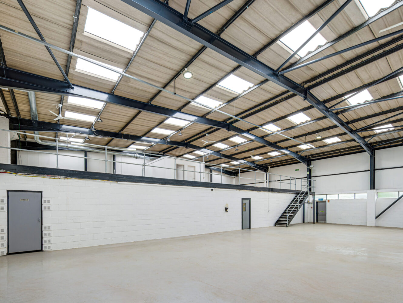 A spacious, empty industrial warehouse with a high ceiling, white brick walls, large windows, and a metal staircase leading to a mezzanine level.