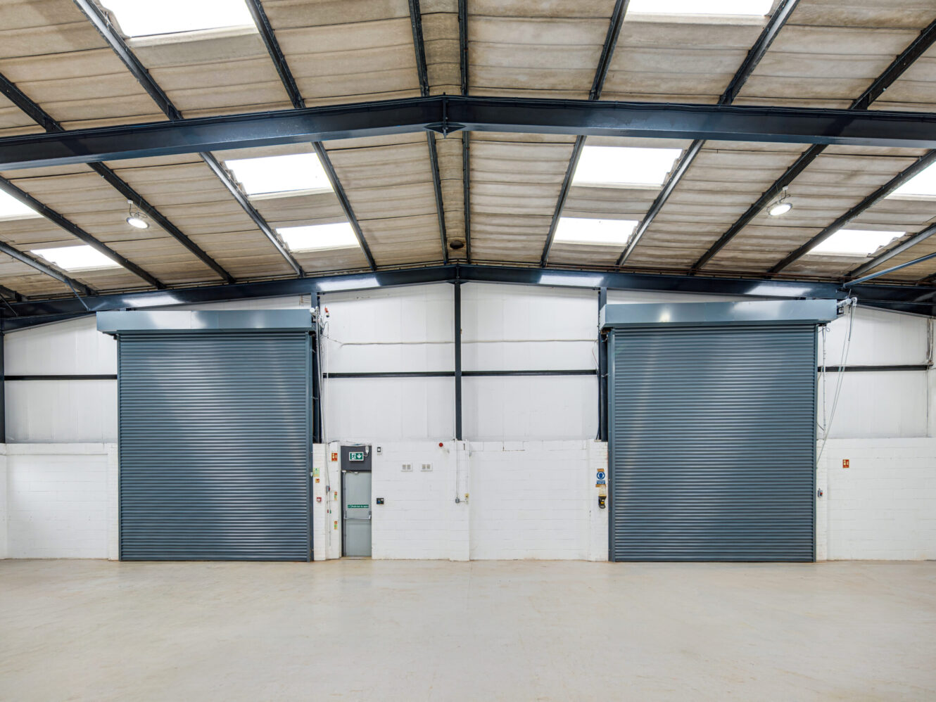 Interior of an empty industrial warehouse with two large metal roller shutter doors, white walls, and a high ceiling with exposed beams and skylights.